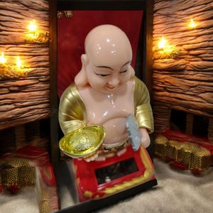 May include: A smiling Buddha figurine with a light pink complexion, gold accents, and a red base. The Buddha holds a gold bowl and a silver object. The figurine is set against a backdrop of wood and candles, creating a warm ambiance.