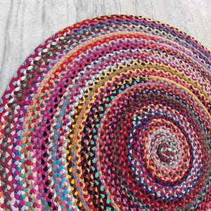 Op de afbeelding: Een rond, gevlochten kleed met een levendig, meerkleurig patroon. Het textiel is gemaakt van strak geweven stoffen stroken in diverse tinten rood, roze, blauw, geel, groen en zwart, gerangschikt in een doorlopend, concentrisch spiraalontwerp.