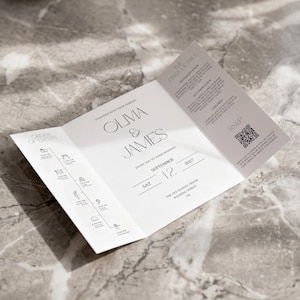 May include: A white wedding invitation, folded into three sections, with the names "Olma & James" in elegant script. The invitation includes the date, September 12, 2027, and details about the wedding. The background is a marble surface.