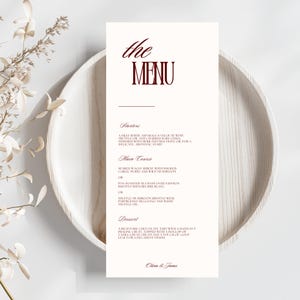 May include: A white menu card with the words "the MENU" in burgundy script. The menu lists starters, main courses, and dessert options. The card is on a light wooden plate, with dried flowers in the background.