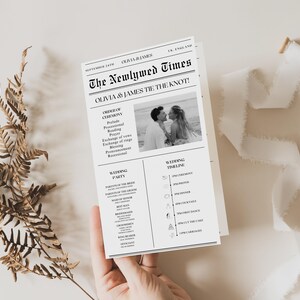 May include: A wedding program designed to look like a newspaper, titled "The Newlywed Times." The black and white design includes a photo of the couple, wedding details, and a timeline. A fern and ribbon are in the background.