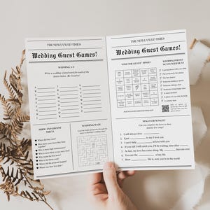 Op de afbeelding: Twee vellen met huwelijksgastspellen met de kop "Wedding Guest Games!" in een lettertype in krantenstijl. De spellen omvatten een bruiloft A-Z, trivia, een doolhof, bingo en songteksten. De vellen worden door een hand vastgehouden.