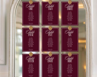 May include: Nine burgundy table cards with gold accents are displayed on a mirror. Each card has a gold wax seal and white text, listing table numbers and guest names. The cards are arranged in a grid.
