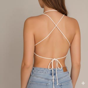 May include: Cream-colored halter top with a crisscross back design. The top features a halter neck and ties at the lower back. The garment is paired with light blue denim jeans.