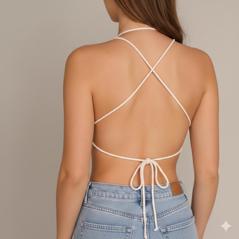 May include: Cream-colored halter top with a crisscross back design. The top features a halter neck and ties at the lower back. The garment is paired with light blue denim jeans.
