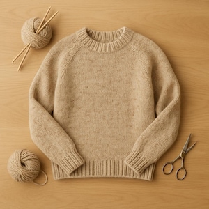 May include: A beige knit sweater with a crew neck and ribbed cuffs and hem. Two balls of yarn, knitting needles, and scissors are arranged around the sweater on a light wood surface. The sweater is a neutral color.