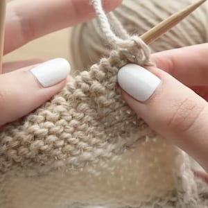 May include: Close-up of hands knitting with beige yarn and wooden needles. The knitted fabric shows a textured pattern with light and dark beige stripes. A ball of yarn is visible in the background, suggesting a crafting project.
