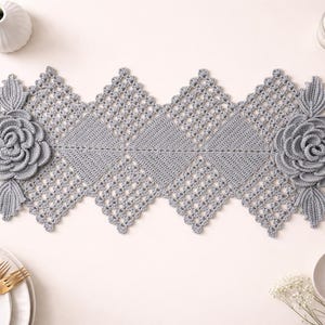 May include: A gray crocheted table runner with a floral design. The runner features a repeating diamond pattern and large, three-dimensional rose appliqués at each end. The runner is approximately 60 inches long.