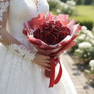 Puede incluir: Un ramo de rosas rojo oscuro envuelto en papel rojo y blanco, atado con una cinta roja. El ramo se sostiene frente a un vestido de encaje blanco. El fondo presenta un jardín con flores blancas y un camino de piedra.