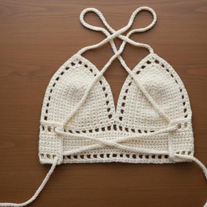 May include: Cream-colored crochet halter top with a crisscross back design. The top features a triangle-shaped bust and adjustable straps. The handmade garment is displayed on a wooden surface, showcasing its intricate stitchwork and openwork detailing.