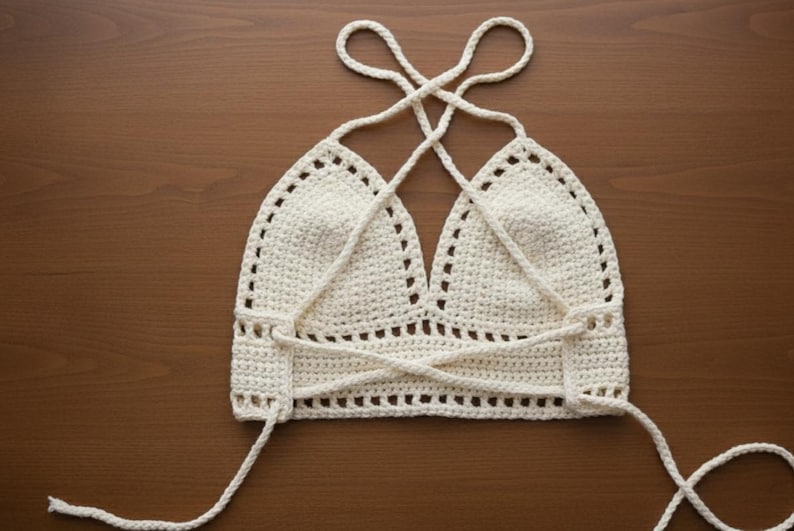 May include: Cream-colored crochet halter top with a crisscross back design. The top features a triangle-shaped bust and adjustable straps. The handmade garment is displayed on a wooden surface, showcasing its intricate stitchwork and openwork detailing.