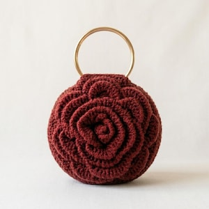 May include: A burgundy crochet handbag with a rose design. The round bag features a gold-colored metal handle. The crochet pattern creates a textured, floral appearance, making it a unique accessory.