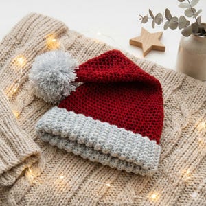 Könnte beinhalten: Ein rot-grauer gestrickter Weihnachtsmannhut mit einem silbernen Bommel, der auf einem beigefarbenen Zopfmusterpullover liegt. Der Hut hat eine graue Krempe und eine rote Krone. Ein Holzstern und eine kleine Vase mit Grünzeug sind im Hintergrund.