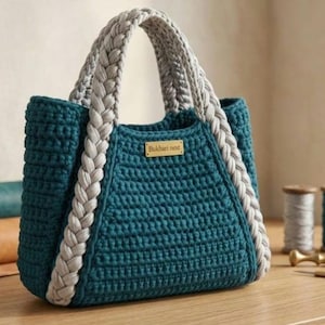 Puede incluir: Bolso tote de ganchillo turquesa con asas y ribetes trenzados grises. El bolso tiene forma rectangular con un patrón de puntadas texturizadas. Una pequeña etiqueta rectangular con el texto "Bukhari nest" está adjunta.