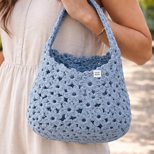 May include: A light blue crochet bag with a floral pattern. The bag has a rounded shape and a sturdy handle. A small white tag with the words "hand made" is sewn on the side. The bag is held by a person.