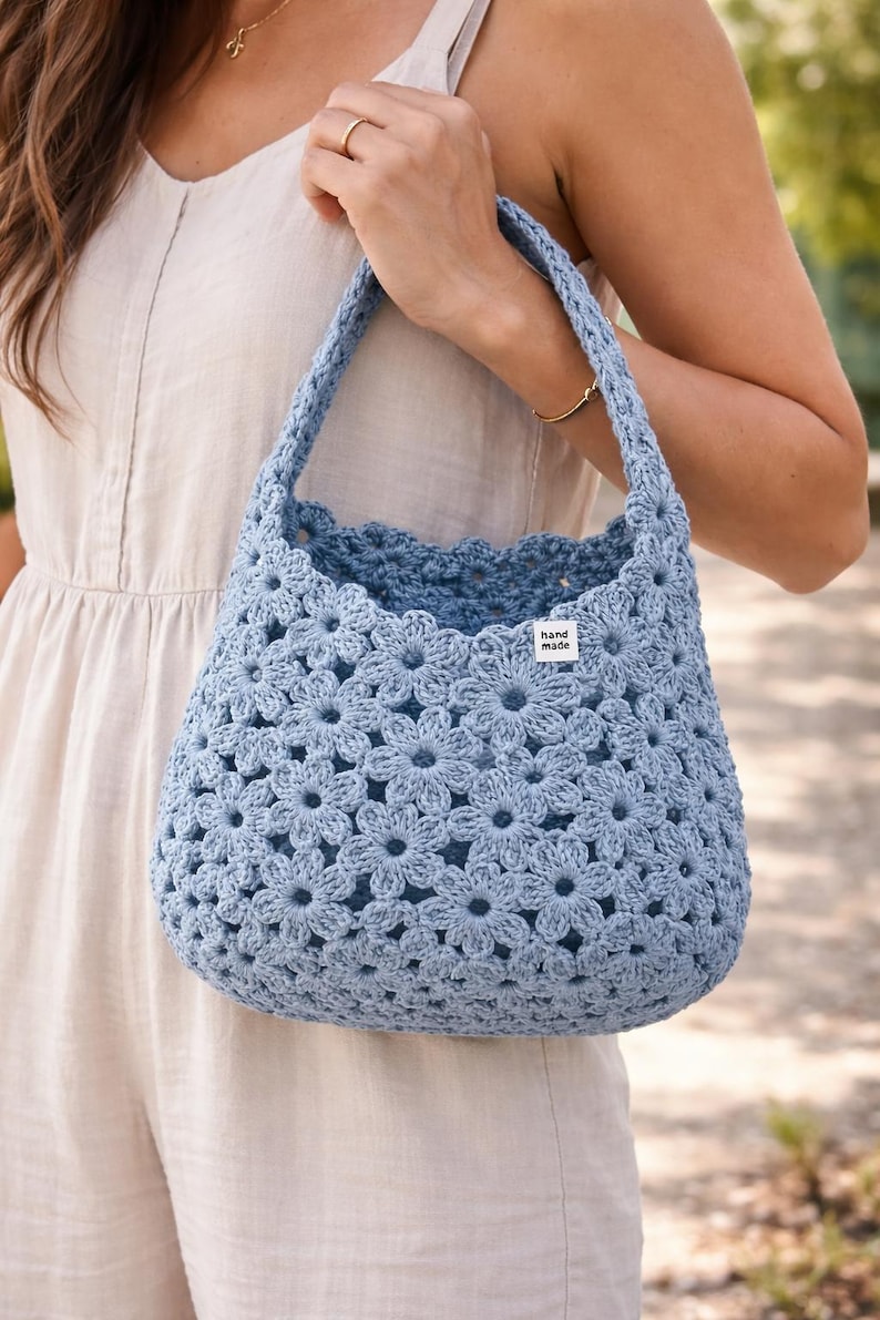 May include: A light blue crochet bag with a floral pattern. The bag has a rounded shape and a sturdy handle. A small white tag with the words "hand made" is sewn on the side. The bag is held by a person.