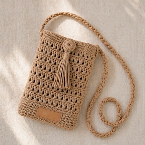 Op de afbeelding: Een bruin gehaakte crossbody tas met een lange, gevlochten riem. De tas heeft een decoratieve kwast en een kleine, rechthoekige patch aan de voorkant. Het haakpatroon heeft driehoekige openingen.