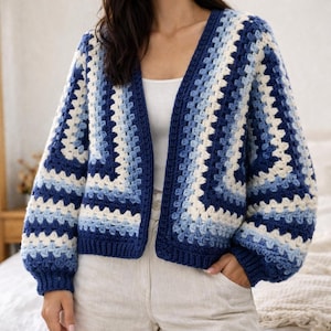 Può includere: Un cardigan fatto a mano, aperto, con un motivo a quadrati granny. Il maglione presenta un design a blocchi di colore con sfumature di blu e bianco. Le maniche sono lunghe e hanno una forma leggermente a sbuffo. Il cardigan è realizzato con un filato morbido e strutturato.