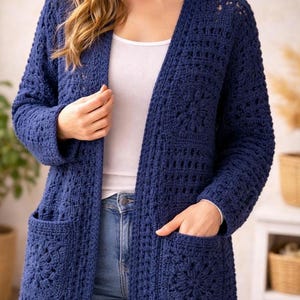 Könnte beinhalten: Marineblauer, offener Häkelcardigan mit zwei Vordertaschen. Der langärmlige Pullover hat ein Blumenmuster auf den Taschen und wird über einem weißen Oberteil und einer blauen Jeans getragen. Der Cardigan ist ein handgefertigtes, gemütliches Kleidungsstück.