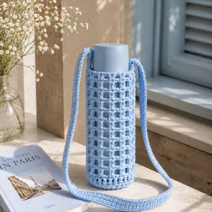 Könnte beinhalten: Hellblaue Wasserflasche in einem gehäkelten Träger mit Riemen. Der Träger hat ein Gittermuster. Ein Buch mit dem Titel "A Month in Paris" liegt neben der Flasche. Die Szene spielt im Freien.