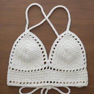 May include: A cream-colored, crocheted halter top with a triangle-shaped bust and a tie closure. The top features a delicate, openwork design along the edges and straps. The straps cross in the back.