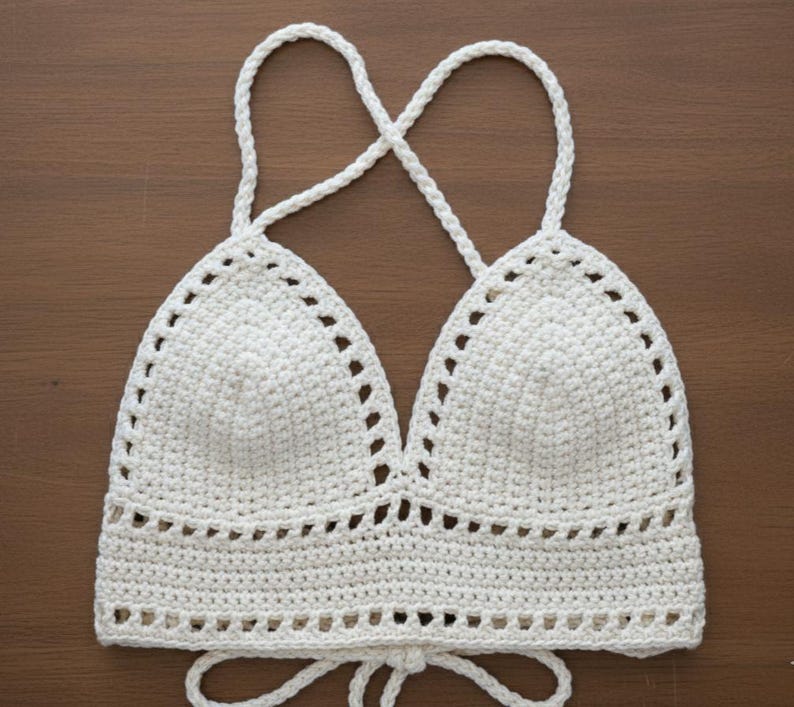 May include: A cream-colored, crocheted halter top with a triangle-shaped bust and a tie closure. The top features a delicate, openwork design along the edges and straps. The straps cross in the back.