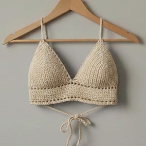 May include: Cream-colored crochet top with a triangle-shaped bust and a tie closure. The handmade top is displayed on a wooden hanger against a neutral background. The top features a textured stitch pattern.