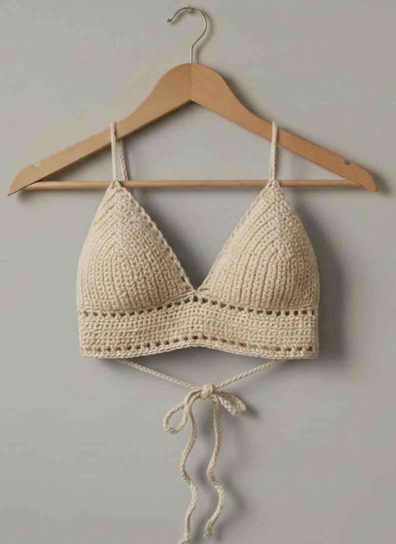 May include: Cream-colored crochet top with a triangle-shaped bust and a tie closure. The handmade top is displayed on a wooden hanger against a neutral background. The top features a textured stitch pattern.