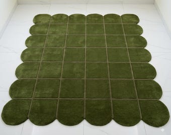Puede incluir: Una alfombra verde con un diseño de borde festoneado único. La alfombra está dividida en secciones cuadradas con bordes verdes más oscuros. El color general es un verde oliva apagado, creando un aspecto suave y acogedor. La alfombra está colocada sobre un suelo de baldosas blancas.