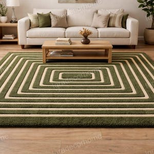 Può includere: Un tappeto rettangolare verde e crema con un motivo geometrico quadrato. Il tappeto è posato su un pavimento in legno in un soggiorno. Il design del tappeto presenta quadrati concentrici, creando un'estetica moderna.