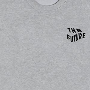 Puede incluir: Una camiseta gris jaspeado con cuello redondo y la frase "THE FUTURE" impresa en negro. El texto est&aacute; ligeramente inclinado y tiene una fuente moderna. La camiseta es de un material suave.
