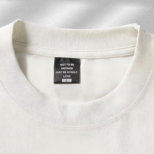 Puede incluir: Primer plano de una camiseta color crema con cuello redondo acanalado. Una etiqueta negra est&aacute; cosida en el cuello, mostrando el texto "NOT TO BE DEFINED JUST BE MYSELF. LIDS" y la talla "L". La camiseta tiene un dise&ntilde;o minimalista.