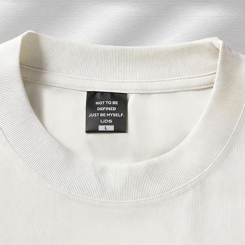 Puede incluir: Primer plano de una camiseta color crema con cuello redondo acanalado. Una etiqueta negra est&aacute; cosida en el cuello, mostrando el texto "NOT TO BE DEFINED JUST BE MYSELF. LIDS" y la talla "L". La camiseta tiene un dise&ntilde;o minimalista.