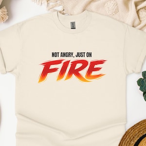 Può includere: T-shirt color crema con la scritta "NOT ANGRY, JUST ON FIRE" in lettere sfumate rosse e arancioni. La maglietta ha un girocollo e maniche corte, adatta per l'abbigliamento casual.