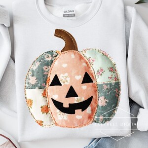 May include: White sweatshirt with a patchwork pumpkin design. The pumpkin features a brown stem, a peach-colored center with white hearts, and floral patterned sections in shades of green and orange. The pumpkin has a black jack-o'-lantern face.