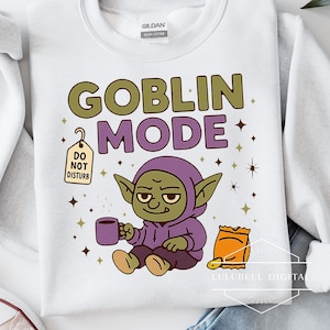 May include: White sweatshirt with the words "GOBLIN MODE" in green and purple. A cartoon goblin in a purple hoodie is holding a mug and a bag of chips. A "DO NOT DISTURB" tag hangs from the design.