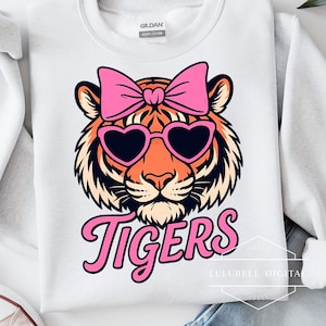 May include: White sweatshirt featuring a tiger face with a pink bow and heart-shaped sunglasses. The word "Tigers" is written in pink script below the tiger's face. The sweatshirt is made of heavy cotton.