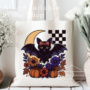 May include: Cream-colored tote bag featuring a black bat with a red bow, set against a crescent moon and a checkered pattern. The design includes pumpkins, flowers, and the text "Also Available in the Shop".