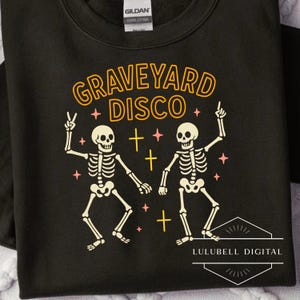 May include: Black sweatshirt with the words "GRAVEYARD DISCO" in orange lettering. Two dancing skeleton figures are featured, along with pink stars and yellow crosses. The sweatshirt has a crew neck and long sleeves.