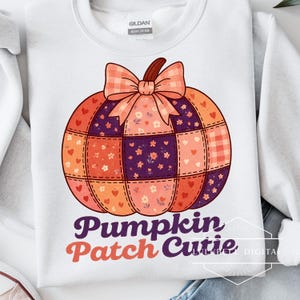 May include: White sweatshirt with a colourful pumpkin design. The pumpkin is made of patchwork squares in orange, purple, and floral patterns, topped with a plaid bow. The text "Pumpkin Patch Cutie" is printed below the pumpkin.