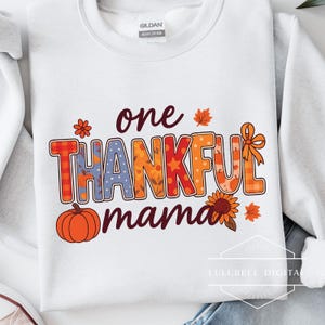 以下が含まれることがあります： 「one THANKFUL mama」の文字が秋をテーマにしたカラフルな文字でプリントされた白いスウェットシャツ。デザインにはカボチャ、ヒマワリ、紅葉が含まれています。スウェットシャツはヘビーコットン製です。