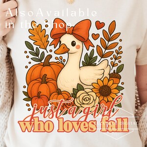 May include: Cream-colored t-shirt featuring a cartoon duck with an orange bow, surrounded by pumpkins, sunflowers, and fall foliage. The text "Just a girl who loves fall" is displayed in a stylized font.