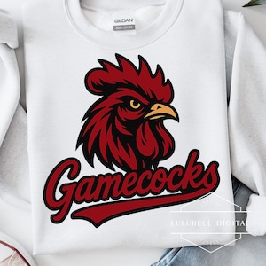 Może przedstawiać: Biała bluza z czerwono-czarnym kogutem i napisem "Gamecocks" w ozdobnej czcionce. Projekt koguta zawiera żółty dziób i oko.