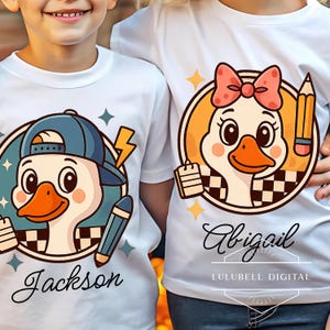 Pode incluir: Camisetas brancas com desenhos de patos de desenho animado. Um pato usa um boné azul e segura um lápis, com o nome "Jackson" abaixo. O outro pato tem um laço rosa e um lápis, com o nome "Abigail". As camisetas são da LULUBELL DIGITAL.