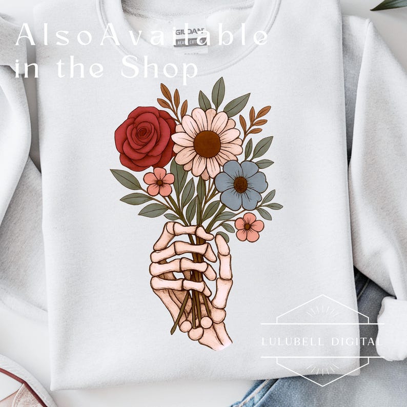 May include: White sweatshirt featuring a graphic of a skeleton hand holding a bouquet of flowers. The bouquet includes a red rose, pink and white daisies, and blue and pink accent flowers. The text "Also Available in the Shop" is at the top.