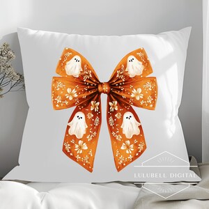 May include: White square pillow featuring a large orange bow design with white ghost illustrations and floral accents. The bow is centered on the pillow, creating a festive Halloween theme. The pillow is on a white surface.