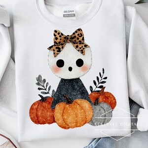 Pode incluir: Camisola branca com uma ilustração de uma figura fantasmagórica com um laço de padrão de leopardo, rodeada por abóboras e folhagem. O design tem um tema de outono ou Halloween.