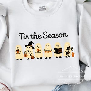 Könnte beinhalten: Weißes Sweatshirt mit dem Text "Tis the Season" in schwarzer Schrift. Darunter Cartoon-Geister mit Halloween-Accessoires, wie einem Hexenhut, einem Besen und einer schwarzen Katze. Das Sweatshirt ist ein Gildan Heavy Cotton.