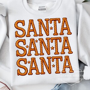 May include: White crewneck sweatshirt with the word "SANTA" in large, rope-style letters in shades of orange and brown. Small star accents are placed within the letters. A festive holiday design.