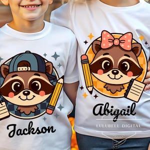 Pode incluir: Duas camisetas brancas com gráficos de guaxinins. Uma camiseta apresenta um guaxinim usando um boné azul, o nome "Jackson" e um lápis. A outra camiseta tem um guaxinim com um laço rosa, o nome "Abigail", um lápis e um caderno.